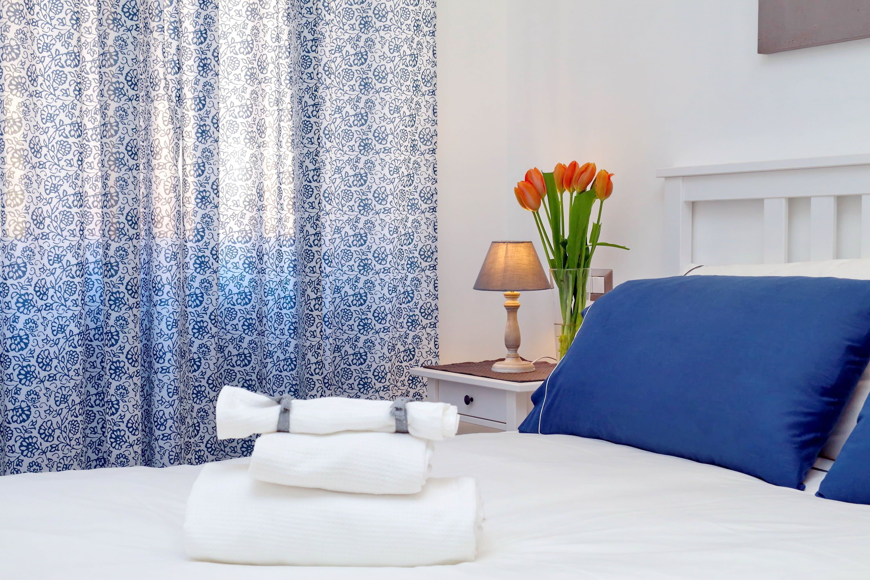 Een netjes opgemaakt bed met blauwe kussens, een vaas met oranje bloemen en een lamp op een nachtkastje.