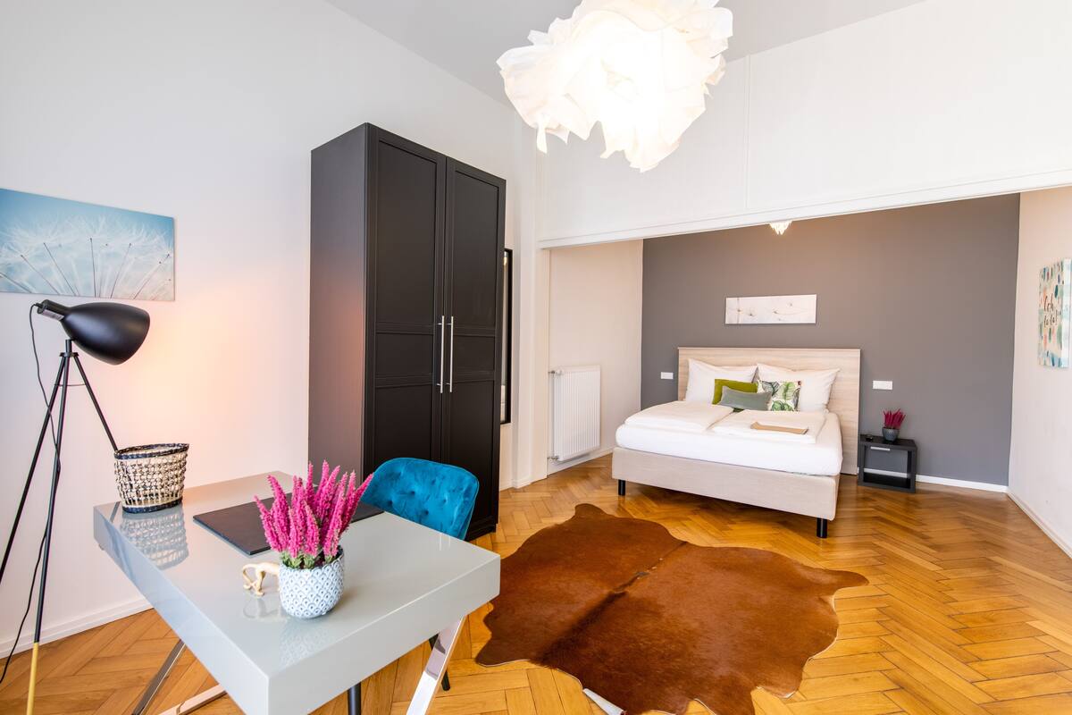 A modern bedroom with a bed, a desk with a lamp, a blue chair, a black wardrobe, and a cowhide rug.