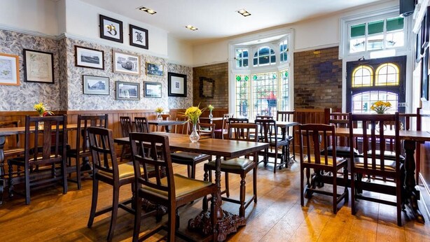 A dining area with wooden tables and chairs, framed pictures on the walls, and a large window with stained glass. - Black Lion (Surbiton)