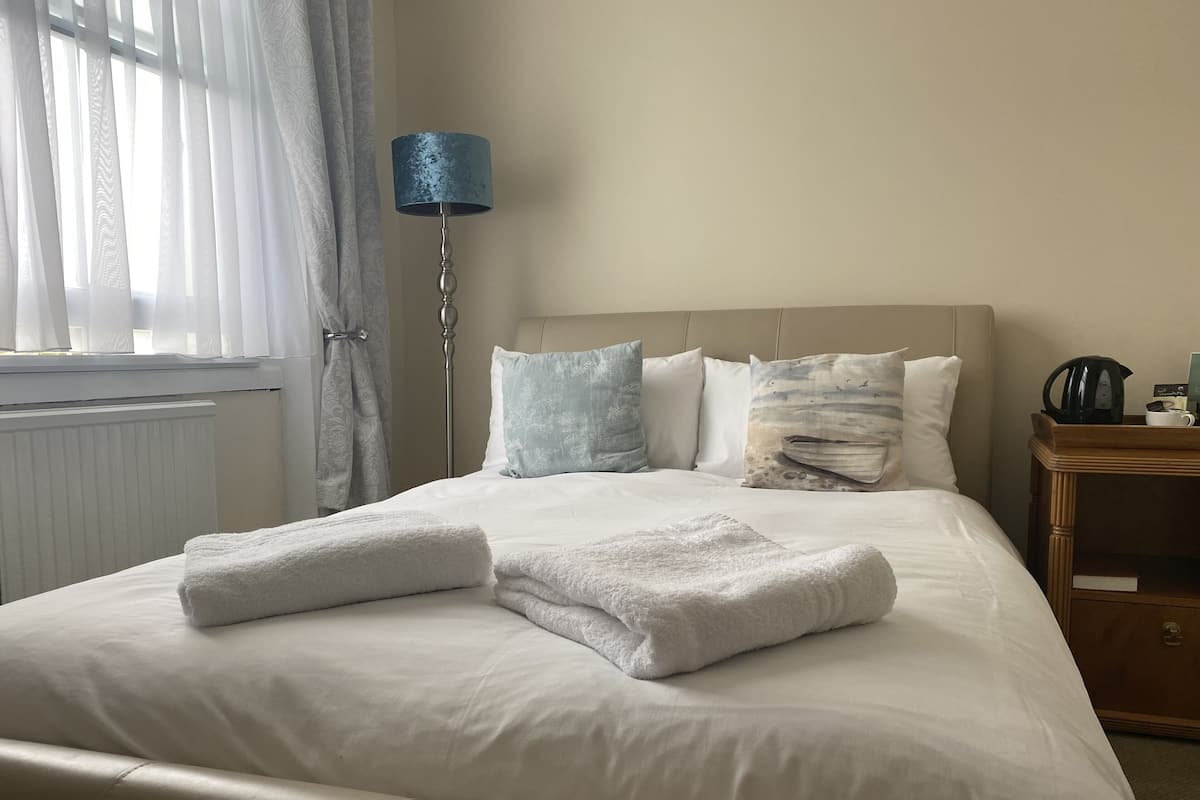 A neatly made bed with white linens and folded towels, a bedside table with a kettle and a cup, and a lamp with a blue shade.