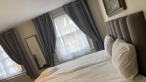 A hotel room with a large bed, grey curtains, a window with sheer curtains, a radiator, and a framed picture on the wall. - Black Lion (Surbiton)