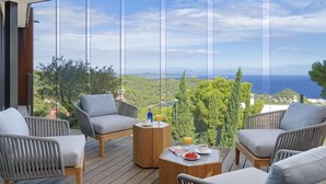 Een balkon met rieten meubels, een houten bijzettafeltje en uitzicht op zee en groen.