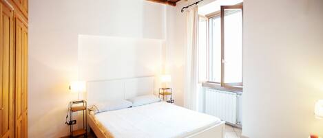 A bedroom with a bed, two bedside tables with lamps, a window with curtains, and a wooden ceiling.