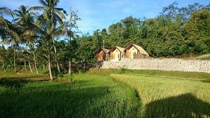A rural setting with thatched-roof cottages, a stone wall, and lush greenery. - Aryasuta Bungalow (Tetebatu)