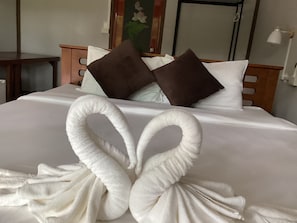 Una cama con una colcha blanca, dos almohadas marrones y dos toallas blancas dispuestas en forma de corazón.