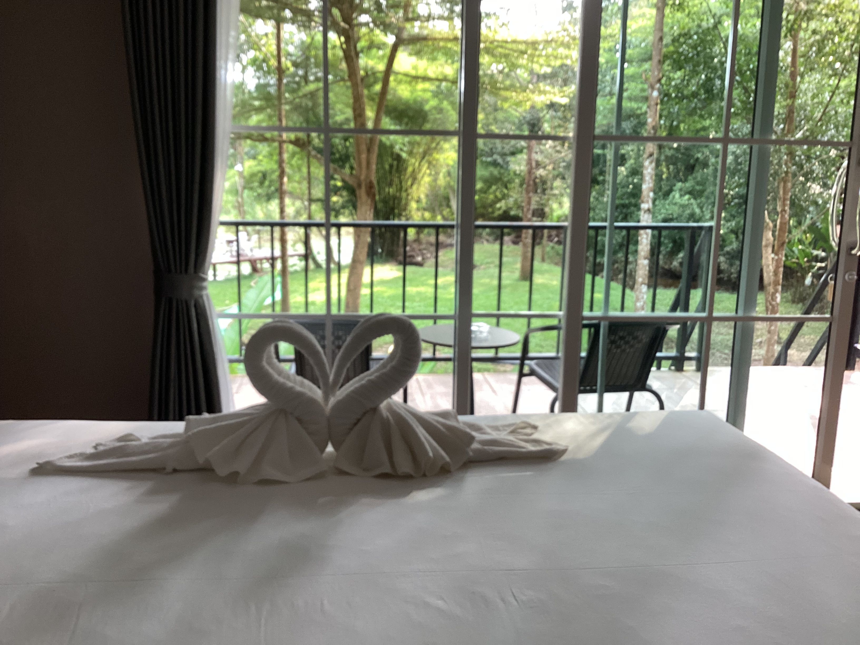 Una cama con una toalla dispuesta en forma de corazón, vistas de un jardín a través de una ventana, y una mesa y sillas afuera.