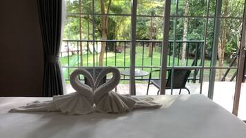 Una cama con una toalla dispuesta en forma de corazón, vistas de un jardín a través de una ventana, y una mesa y sillas afuera.
