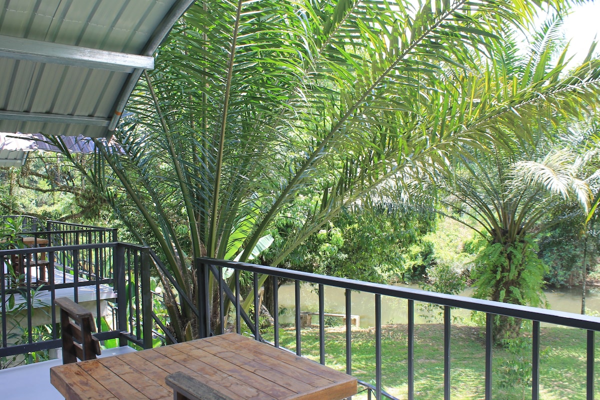 Una mesa y sillas de madera en un balcón con baranda de metal, con vistas a un río y abundante vegetación.