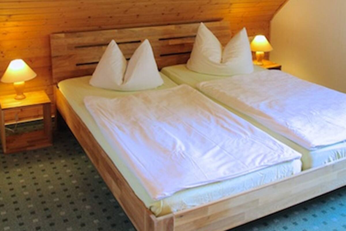 Eine Holzbalkendecke mit geneigter Form, ein Bett mit weißer Bettwäsche und Kissen sowie zwei Nachttischlampen.