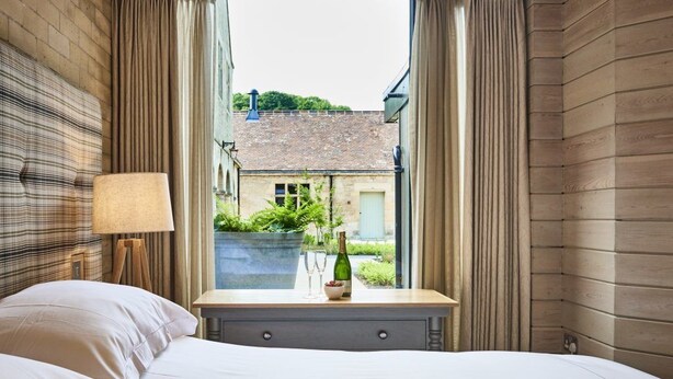 A hotel room with a bed, a bedside table with a bottle and glasses, and a view of a building and greenery outside. - Chesters Stables (Hexham)