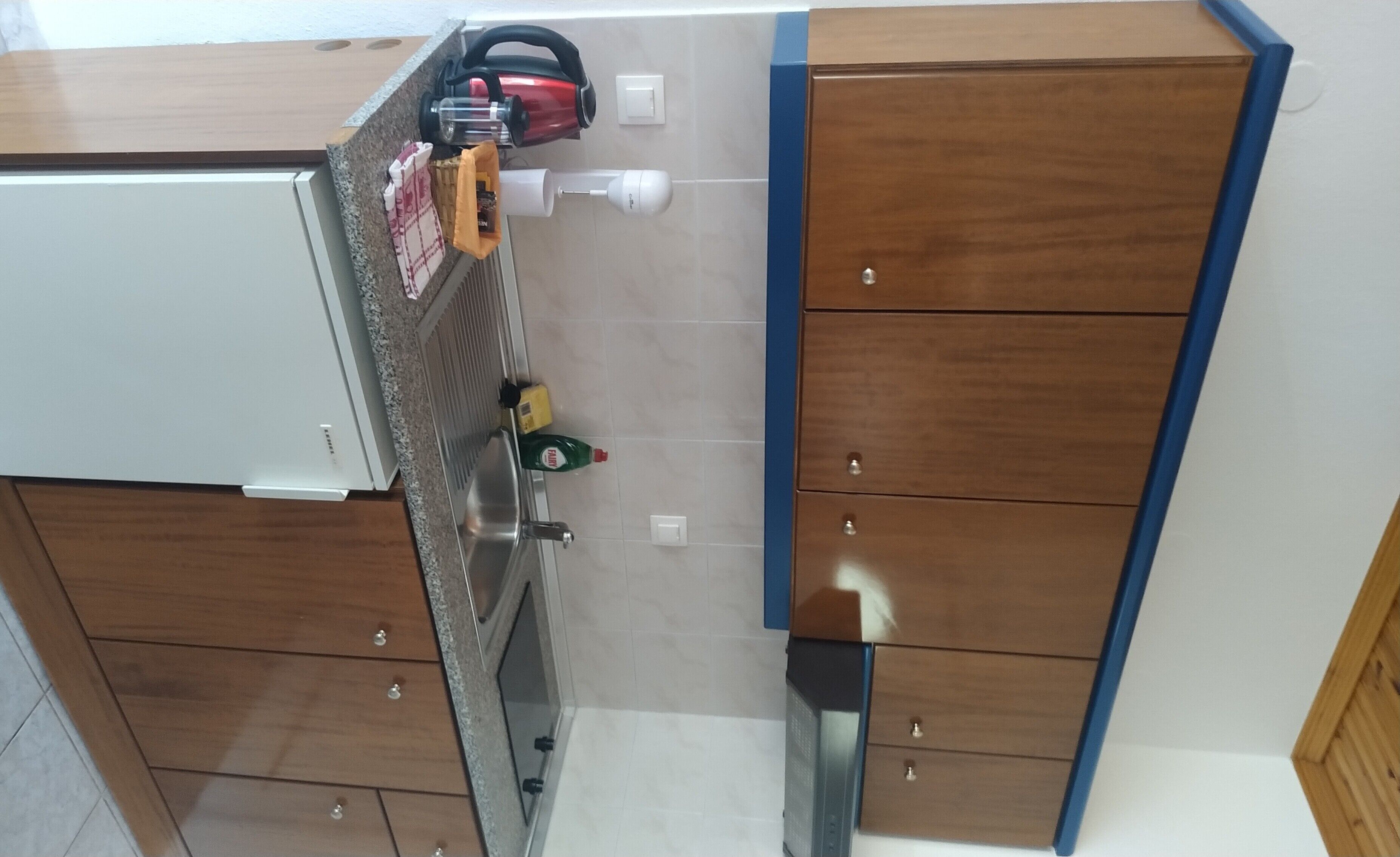 A kitchen with wooden cabinets, a stainless steel sink, and a red kettle on the countertop.