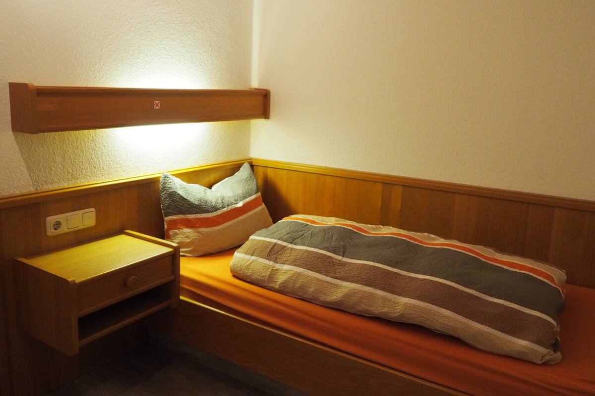 Una habitación pequeña con cama individual, cabecera de madera, mesita de noche con lámpara y toma de corriente.