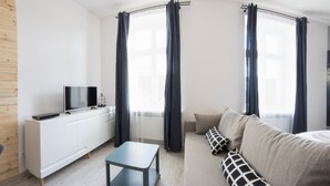 Een moderne woonkamer met een witte kast, een flatscreen-tv, een grijze salontafel, een beige bank met gekleurde kussens en een raam met donkere gordijnen.