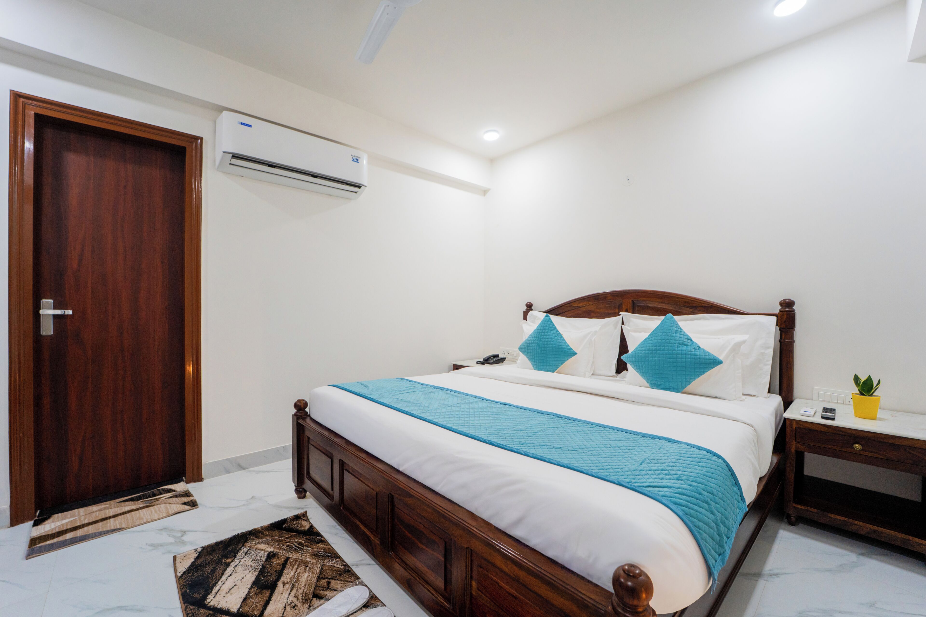 A bedroom with a wooden bed, white bedding, a blue throw, a bedside table with a lamp, and an air conditioning unit.