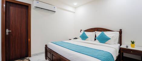 A bedroom with a wooden bed, white bedding, a blue throw, a bedside table with a lamp, and an air conditioning unit.