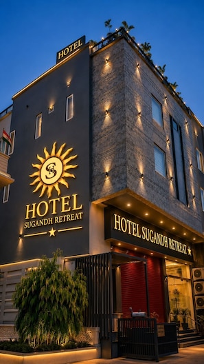 Un hôtel dont la façade est ornée d’un logo représentant un soleil et sur laquelle on peut lire l’inscription « Hotel Sugandh Retreat ».