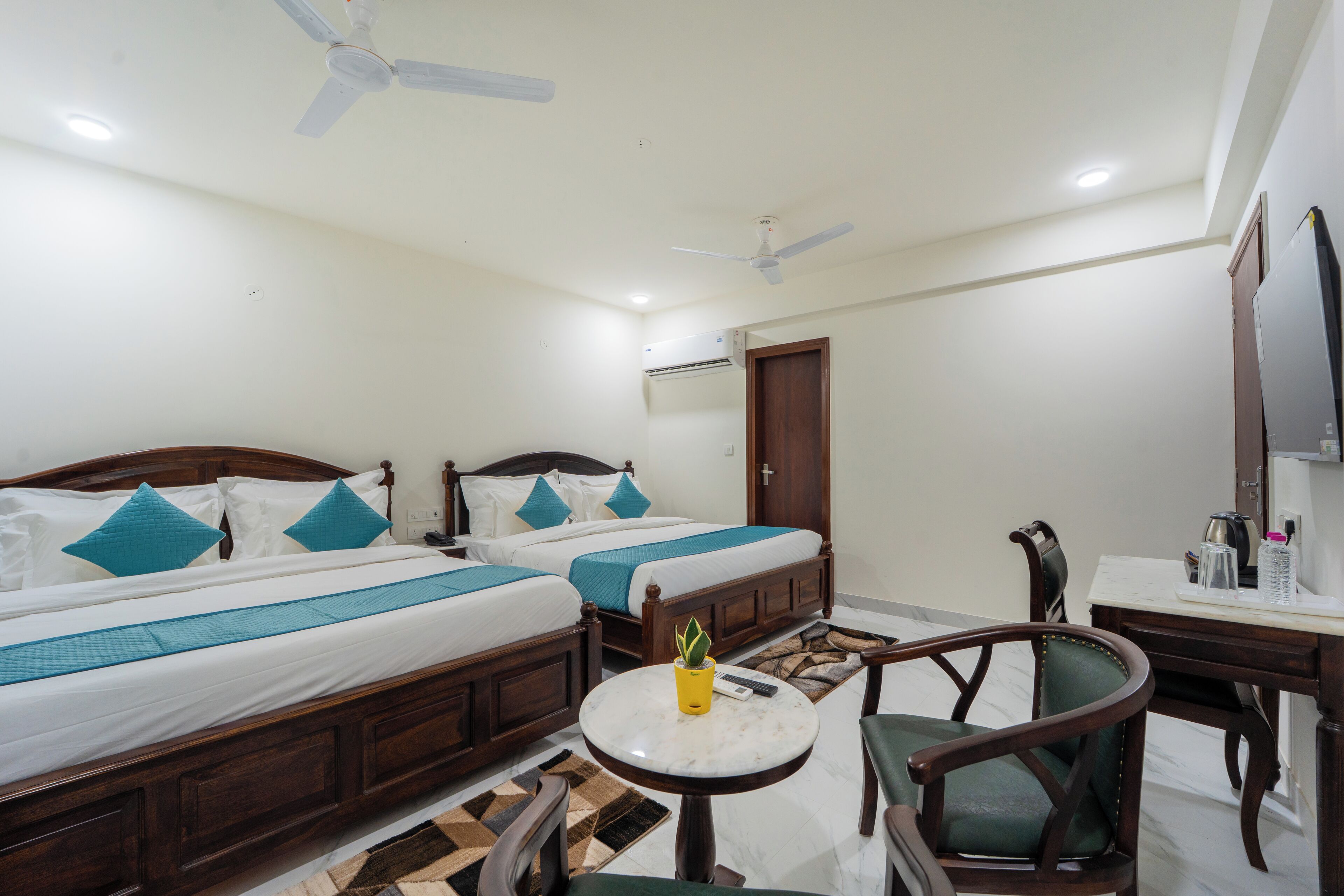 A hotel room with two beds, a small round table, a chair, a desk, a ceiling fan, and an air conditioning unit.