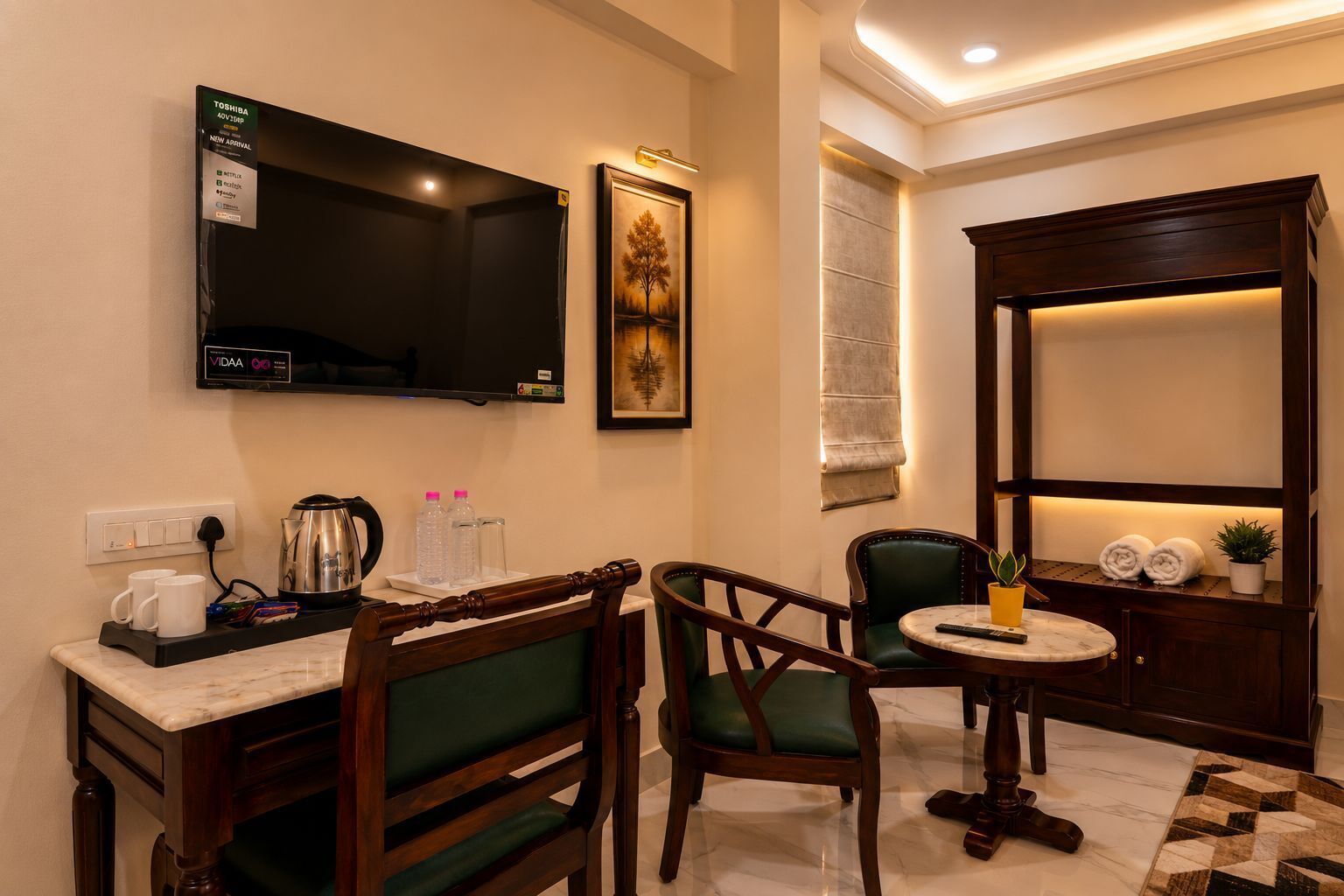 A hotel room with a TV, a painting, a coffee station, a desk, two chairs, a small table, and a cabinet with towels.