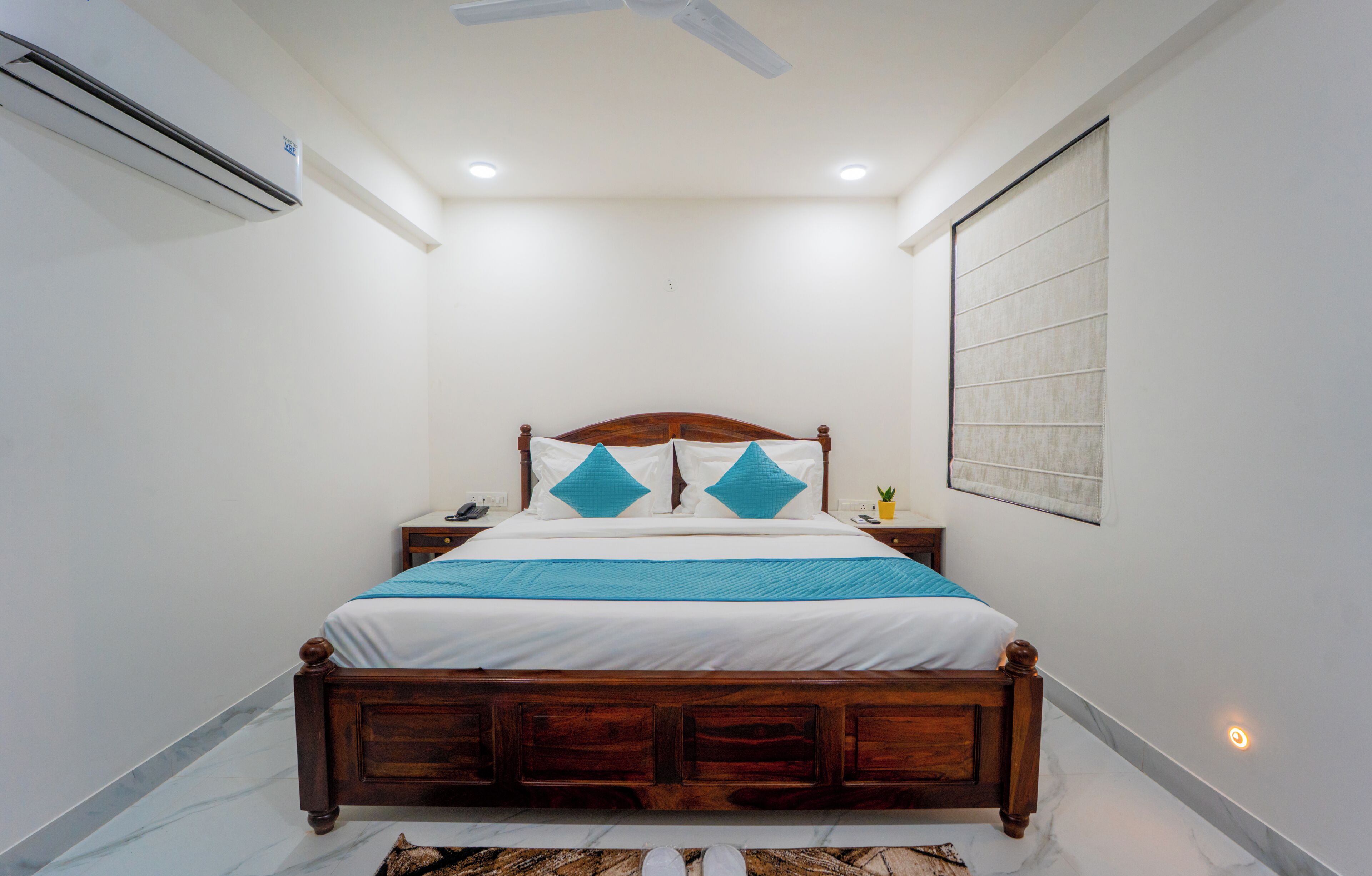 Un dormitorio con cama de madera, paredes blancas, ventilador de techo y mesita de noche con lámpara.