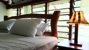 A bed with white bedding and a wooden headboard, a bedside lamp, and a window with blinds. - Sunset Villa Resort (Diani Beach)