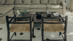 A living room with a sofa, two chairs, a coffee table, and a small table with books and a candle. - Damma Beachfront Luxury Villa (Santorini)