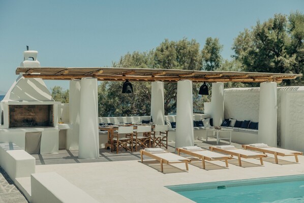 Un área junto a la piscina con una pérgola, mobiliario y una zona de barbacoa.