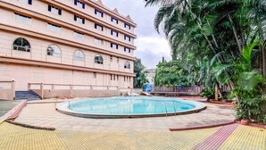 Una piscina con un borde de baldosas, palmeras y un edificio de varios pisos al fondo.