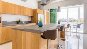 A modern kitchen with wooden cabinets, a central island with bar stools, and large windows offering a view of the city. - Blue Marlin - Famous Handelskade Apartment (Willemstad)