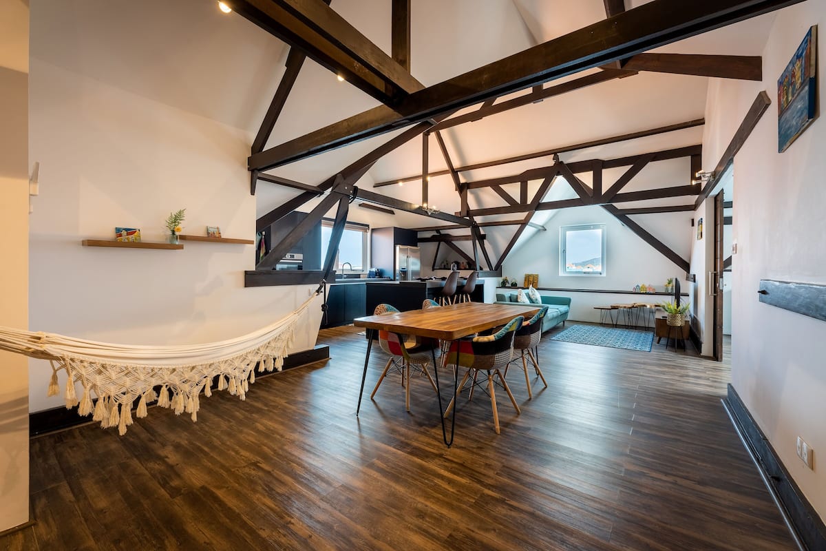 Ein geräumiges Loft mit freiliegenden Holzbalken, einer Hängematte, einem Holzesstisch und einer Küche im Hintergrund.