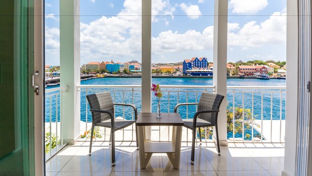A balcony with two wicker chairs and a table, overlooking a marina with colorful buildings and boats. - Blue Marlin - Famous Handelskade Apartment (Willemstad)