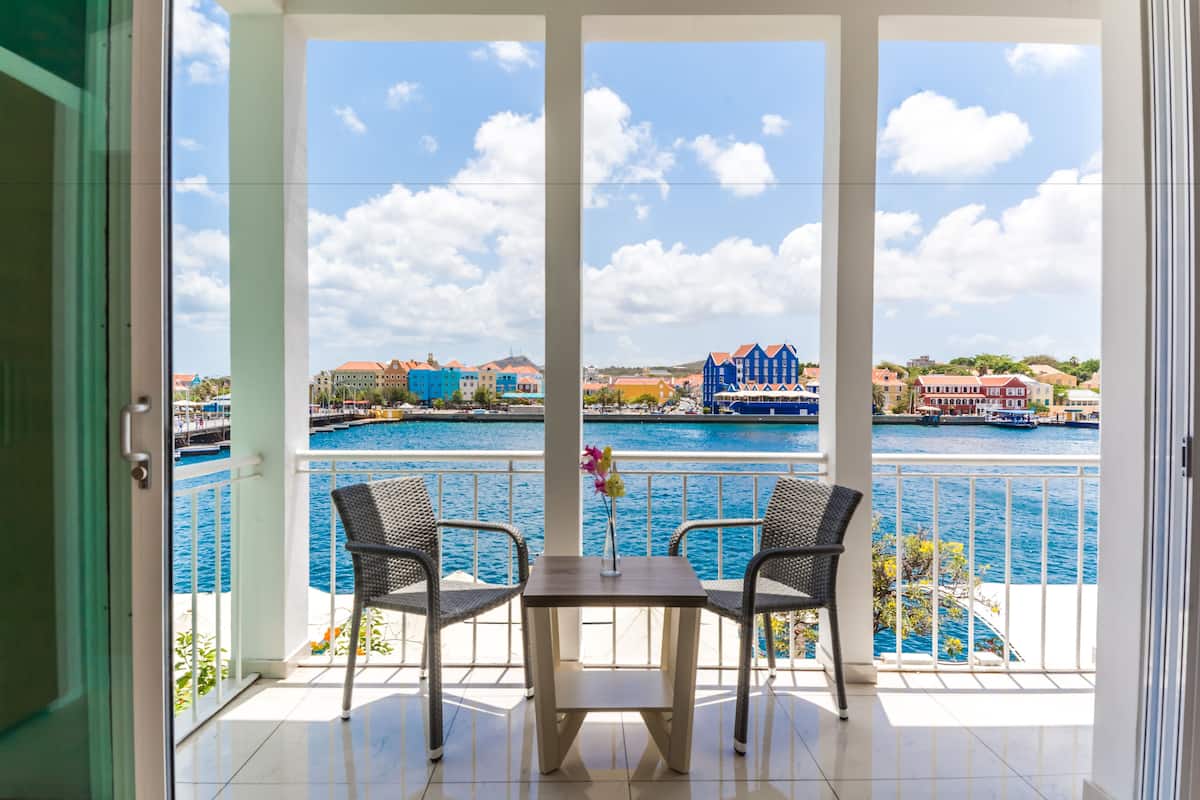 Ein Balkon mit zwei Korbstühlen und einem Tisch, mit Blick auf einen Jachthafen mit bunten Gebäuden und Booten.