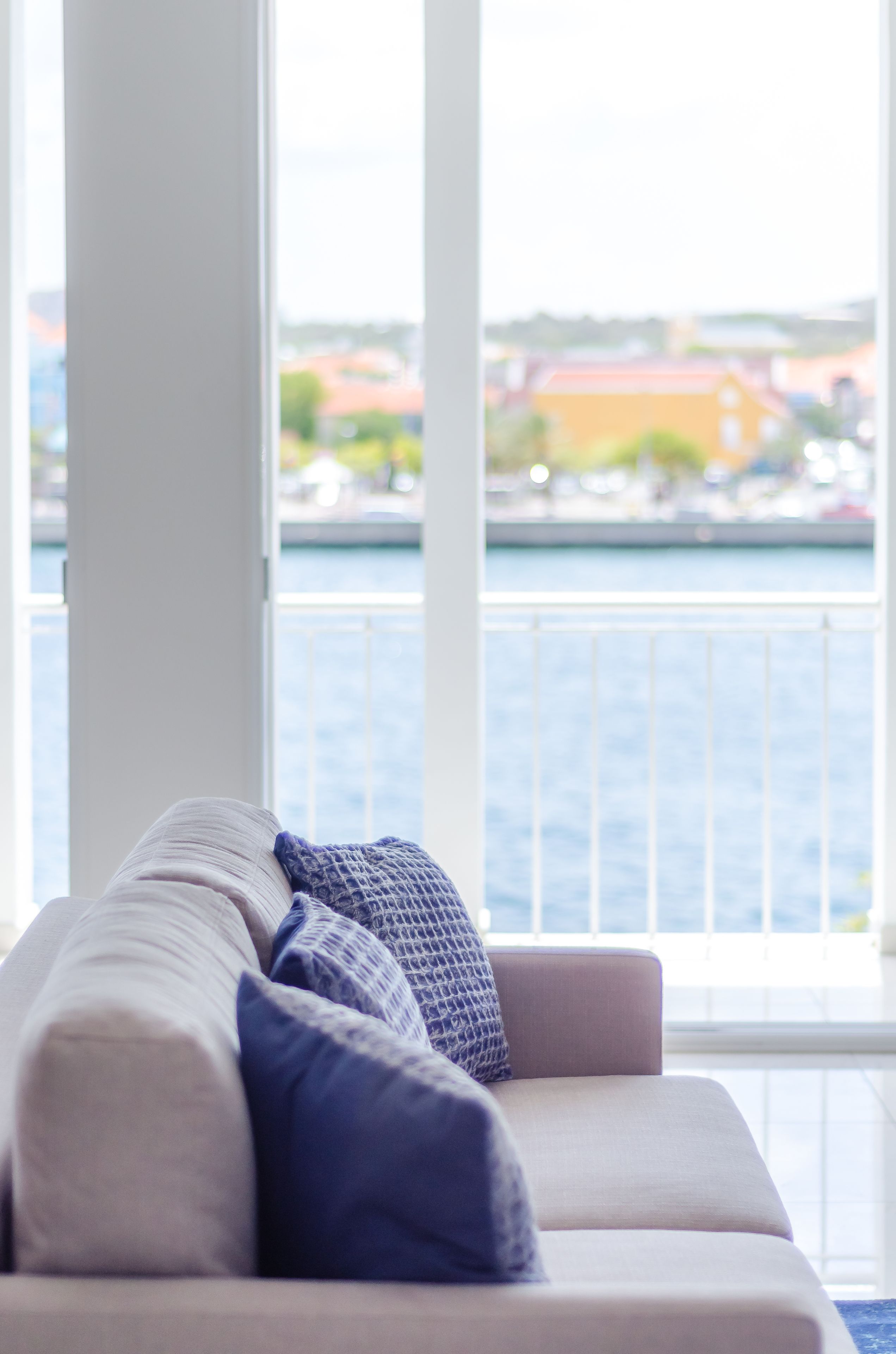 A modern living room with a sofa and large windows overlooking a body of water and buildings.
