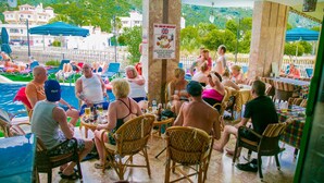 Uma área à beira da piscina com pessoas sentadas em mesas, um letreiro anunciando café da manhã inglês completo e vista da piscina e edifícios ao fundo.