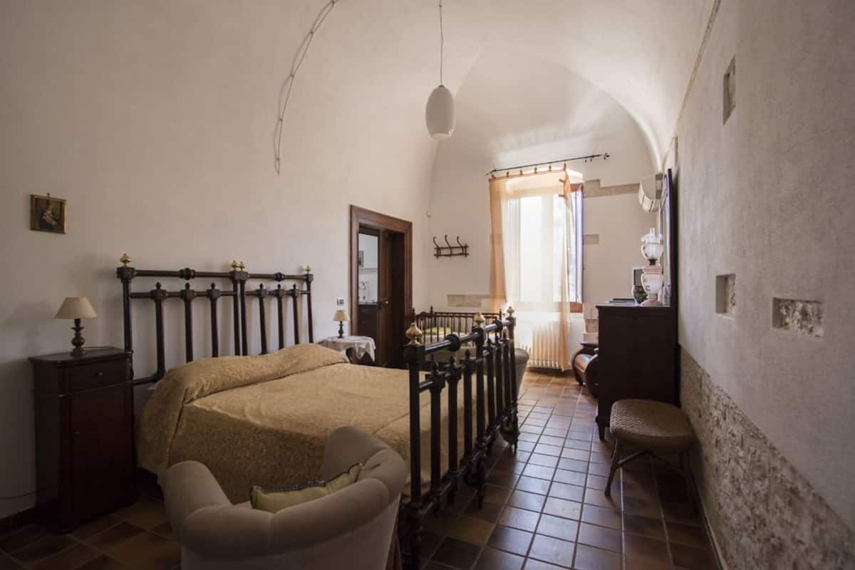 Ein Schlafzimmer mit Bett, Sessel, Nachttisch, Kommode und einem Fenster mit Vorhängen.