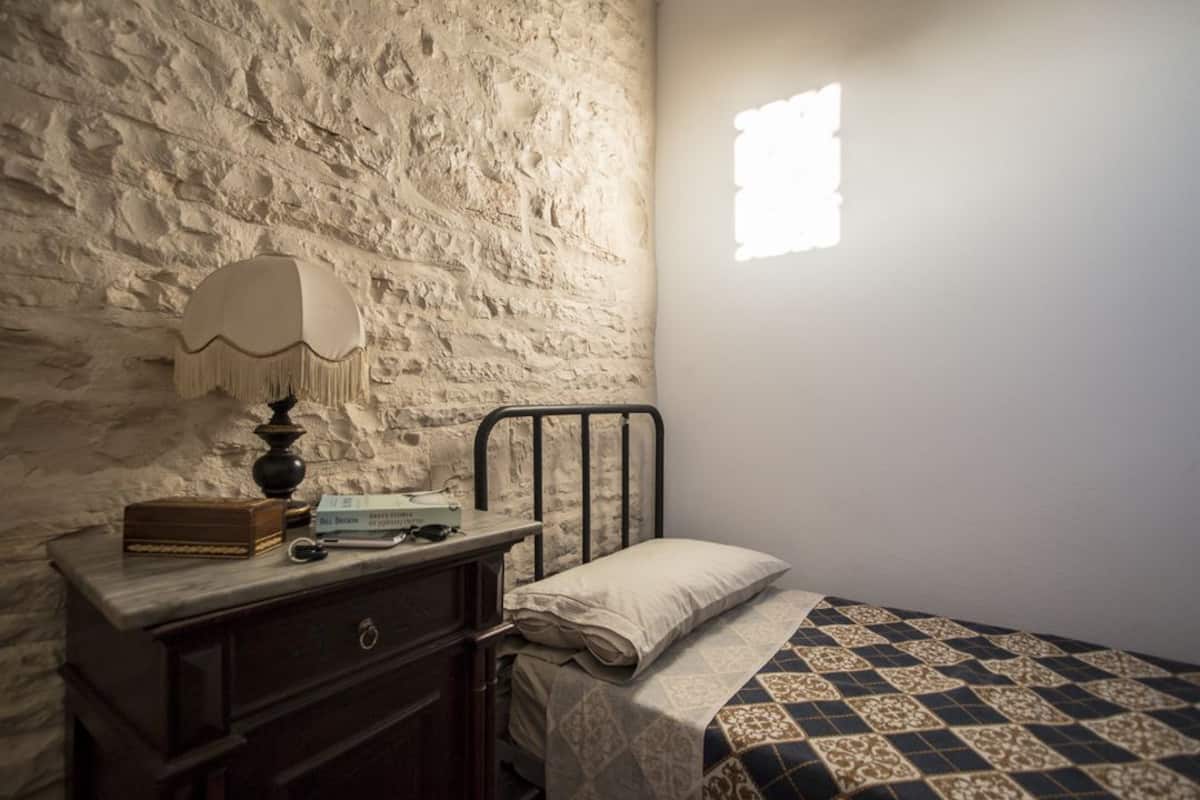 Ein Schlafzimmer mit Steinwand, Nachttisch mit Lampe, Bett mit gemustertem Überwurf und einem Fenster, das Licht an die Wand wirft.