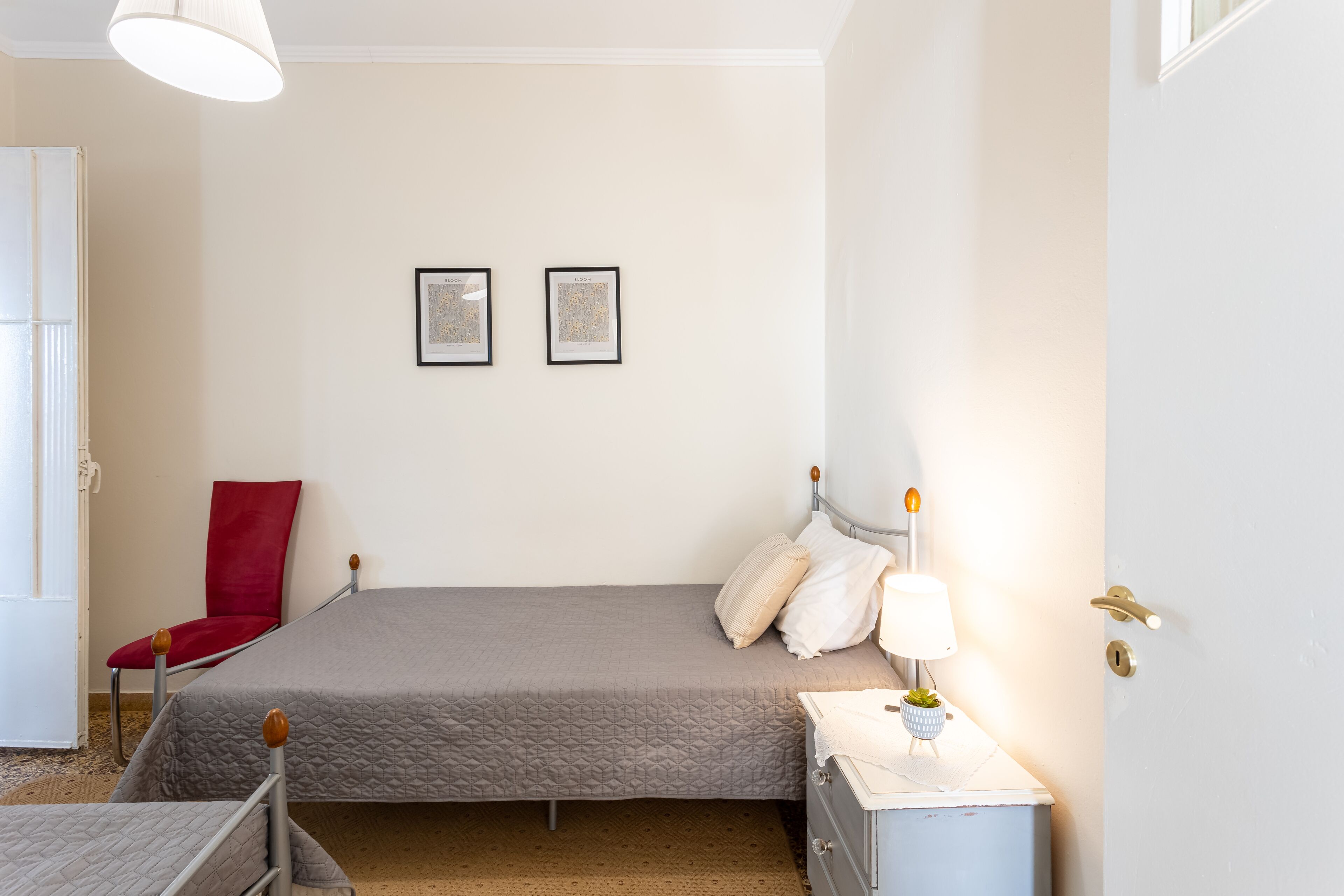 A bedroom with two beds, a red chair, a bedside table with a lamp, and two framed pictures on the wall.
