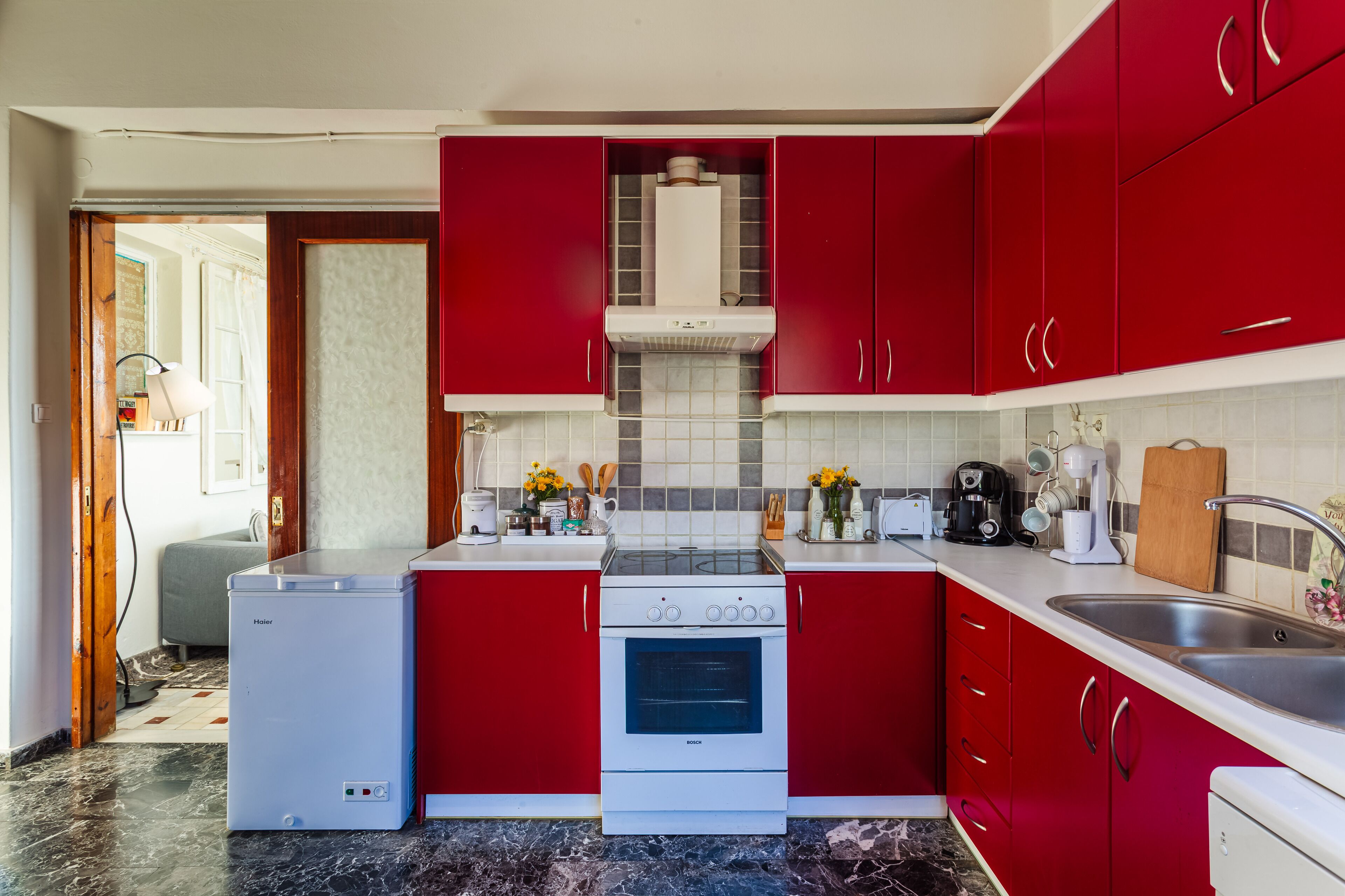 Een kleine keuken met rode kastjes, een Bosch oven, een witte koelkast en een spoelbak.
