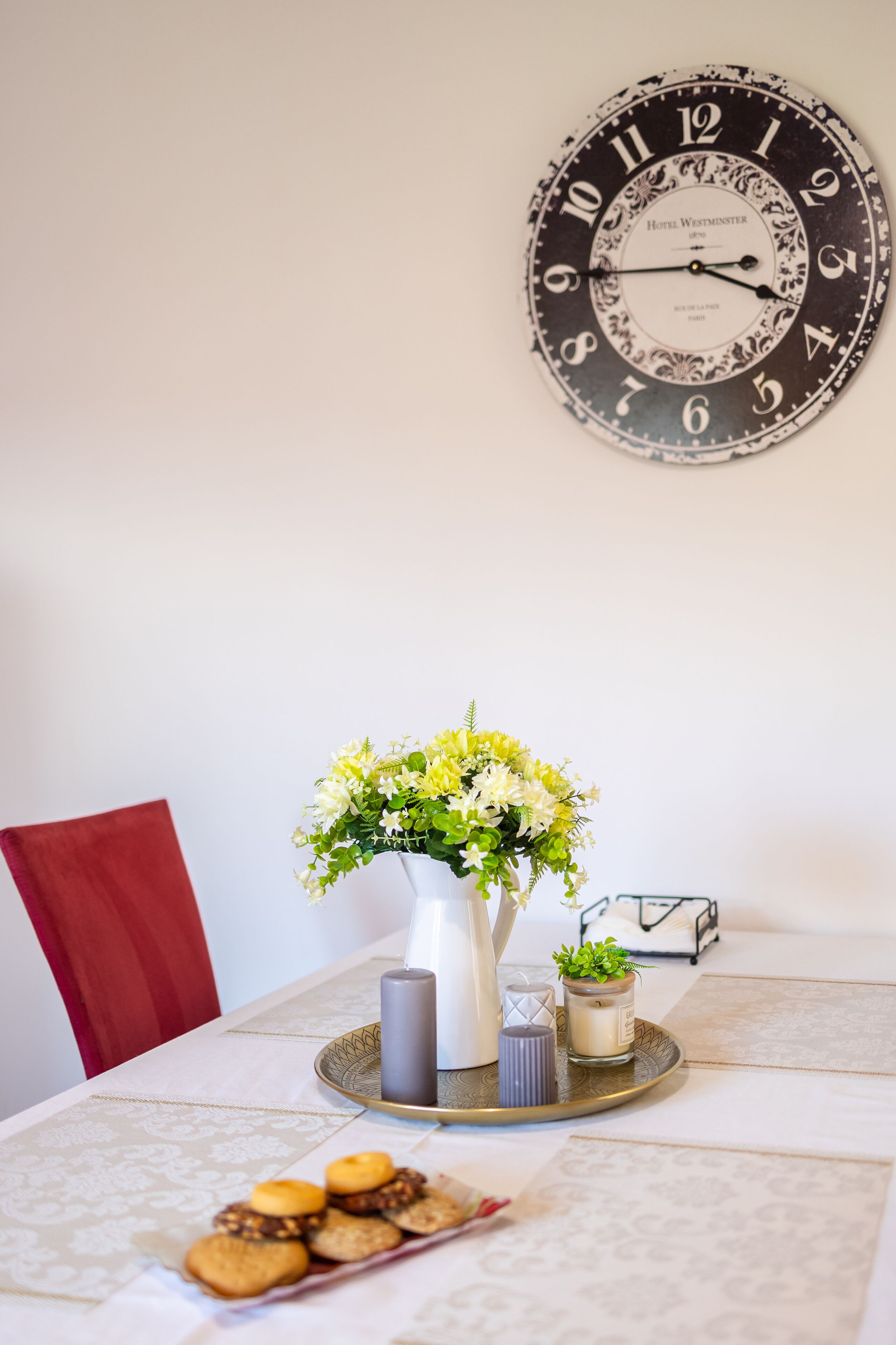 Une table à manger ornée d’une horloge, d’un vase de fleurs, de bougies et d’un plateau de biscuits.