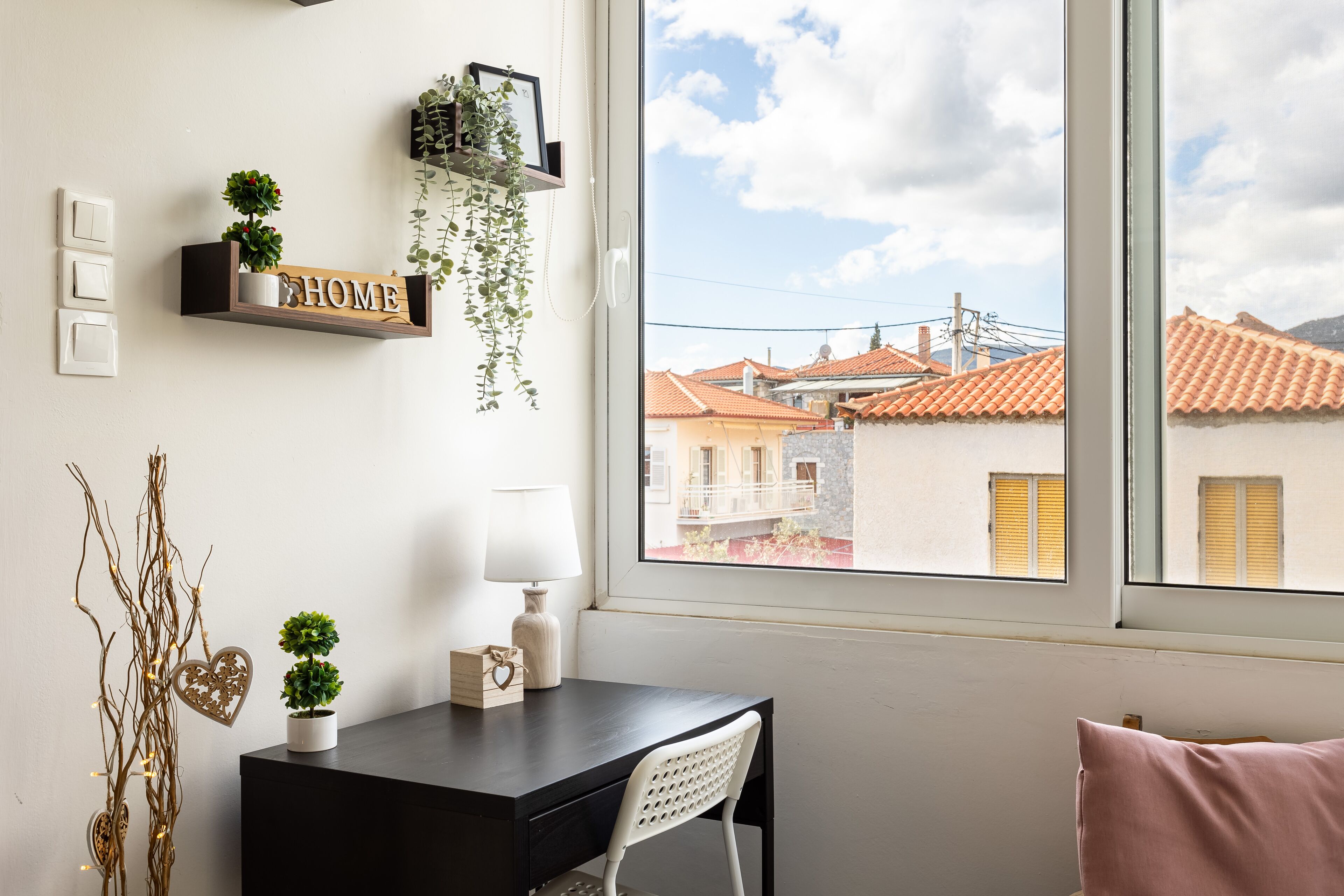 A room with a desk, chair, and a window with a view of buildings. There are wall-mounted lights, a shelf with plants, and a heart-shaped decoration.