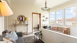 Ein Wohnzimmer mit grauem Sofa, einem kleinen Beistelltisch, einer Chandelierleuchte und Blick durch große Fenster nach draußen.