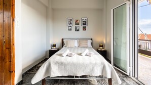 Ein ordentlich eingerichtetes Hotelzimmer mit einem großen Bett, zwei Nachttischen mit Lampen, Blick durch eine Schiebetür und einer Reihe gerahmter Bilder an der Wand.