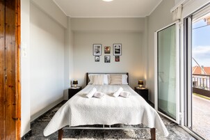 Quarto de hotel bem arrumado, com uma cama grande, duas mesas de cabeceira com luminárias, vista através de uma porta de correr de vidro e uma série de quadros na parede.