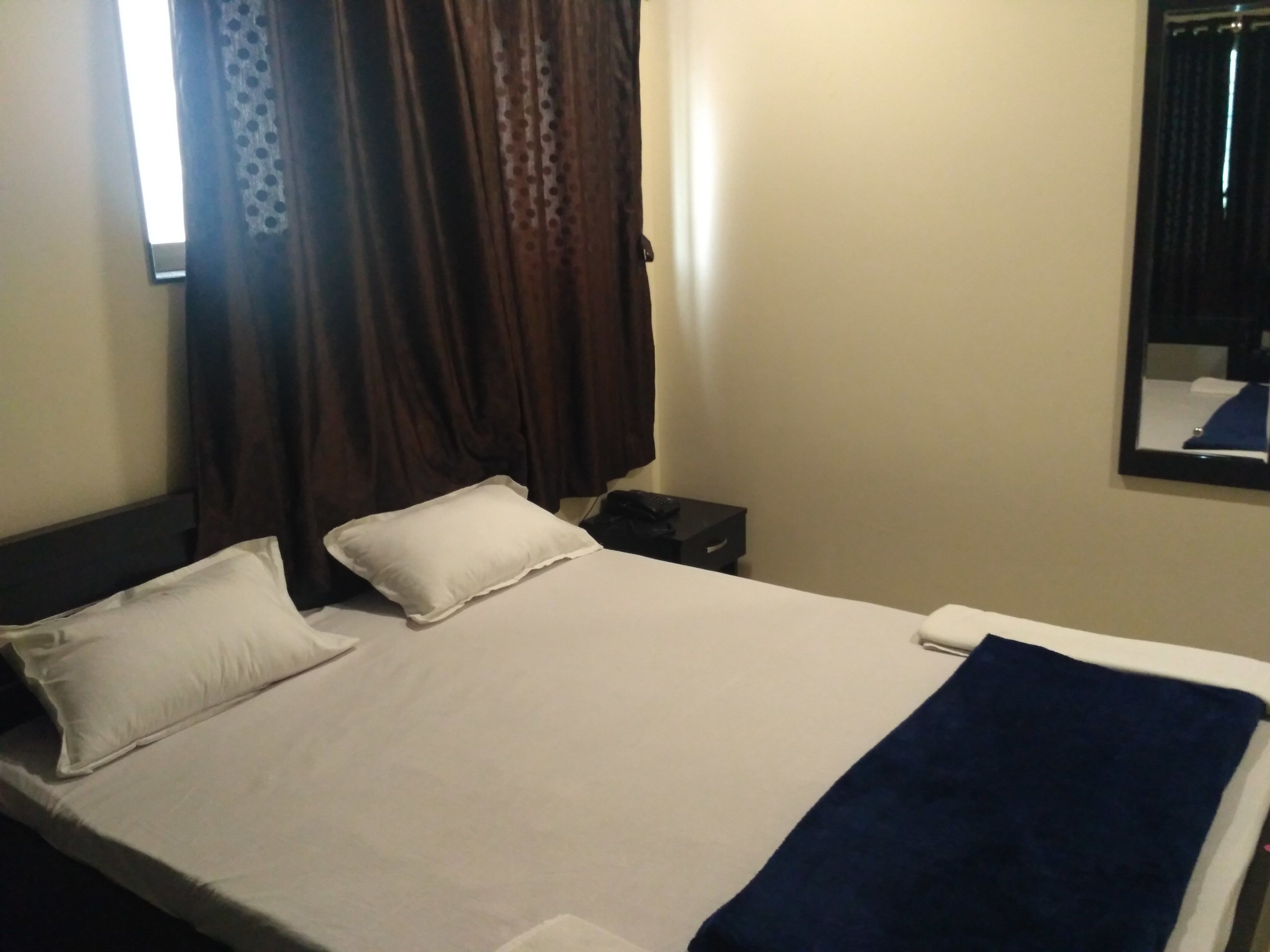 Een hotelkamer met een bed, kussens, dekbed, nachtkastje, spiegel en gordijnen.