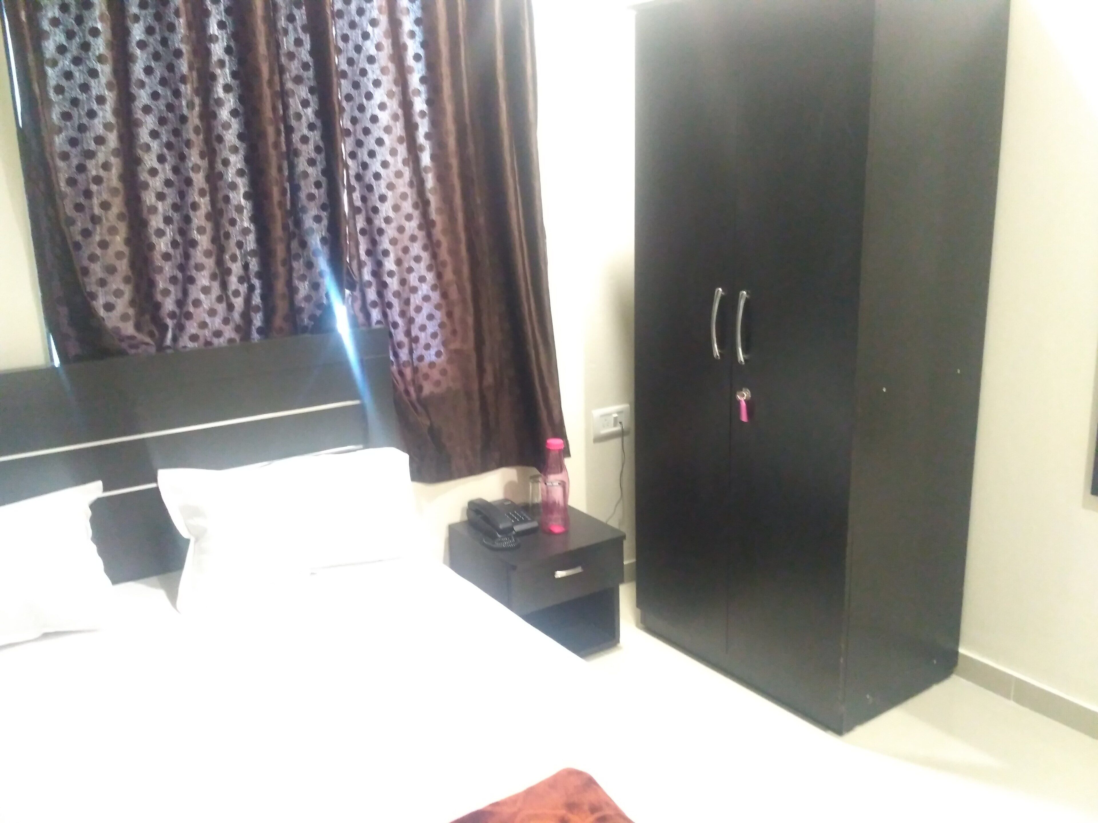 A bedroom with a bed, a wardrobe, a bedside table with a phone and a bottle, and curtains.