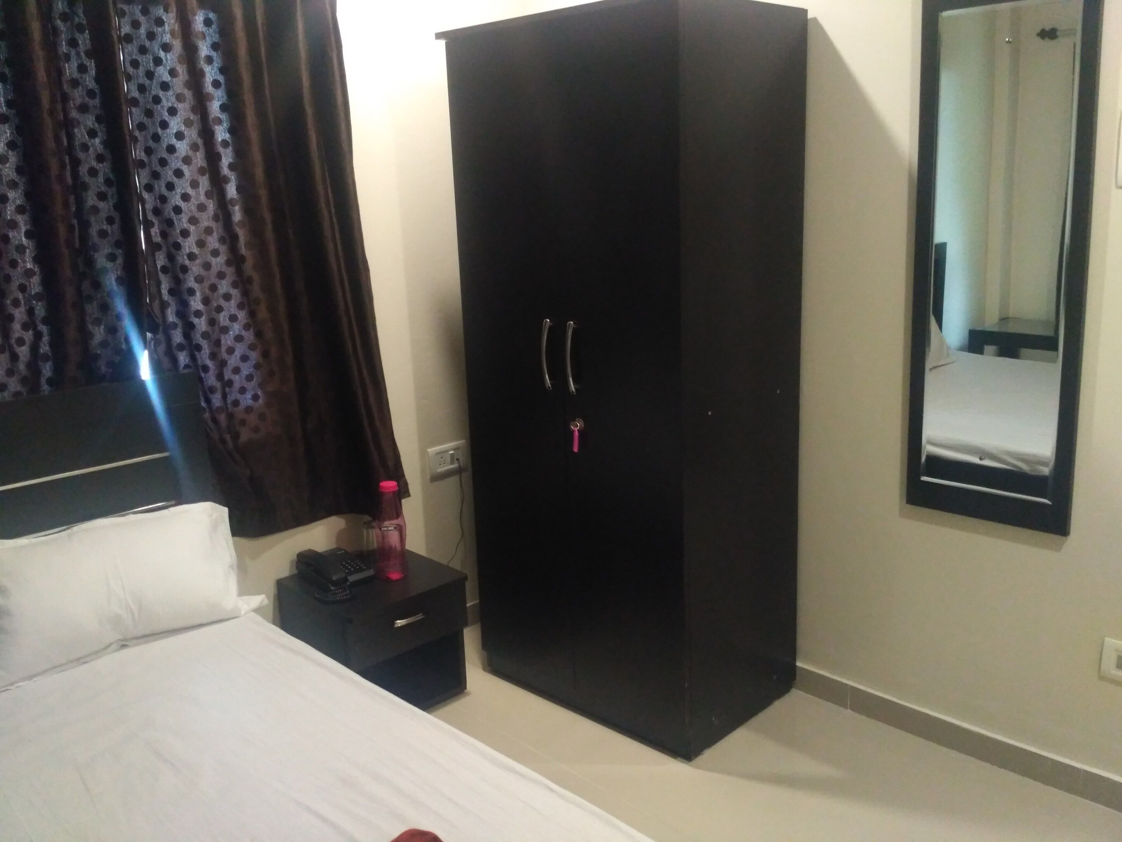 A hotel room with a bed, a black wardrobe, a nightstand with a phone and a bottle, and a mirror on the wall.