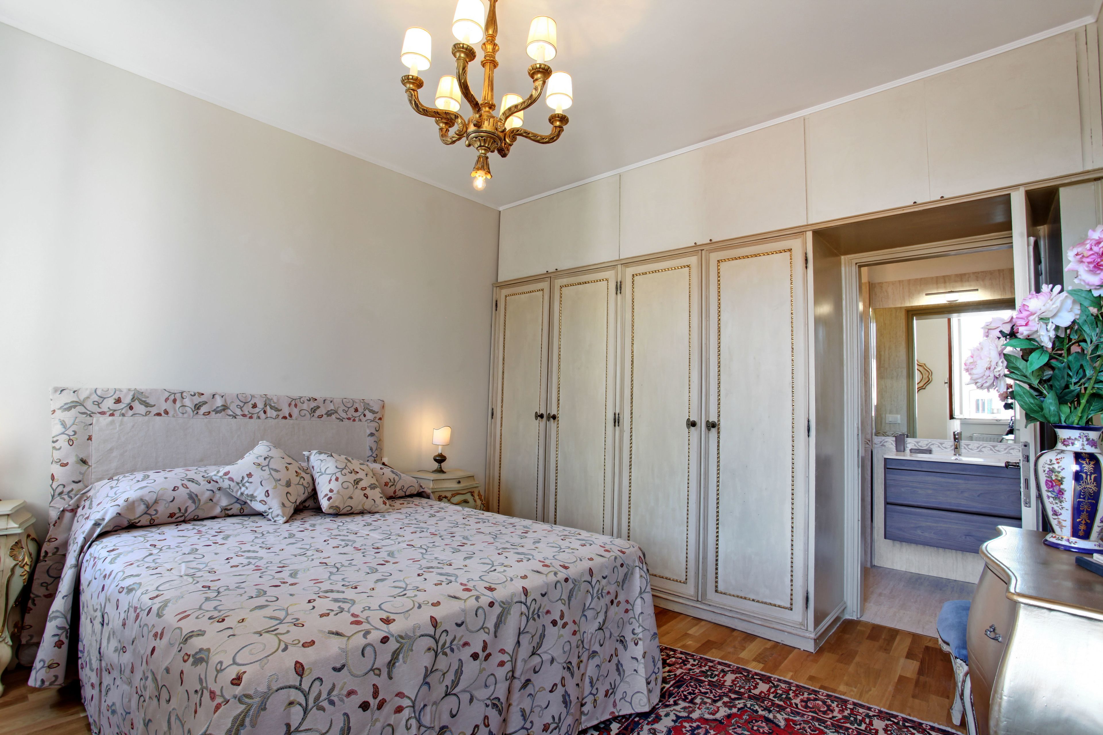 Une chambre à coucher avec un grand lit, un lustre, une armoire et une commode agrémentée d’un vase de fleurs.
