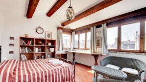 A room with a bed, a wicker chair, a bookshelf, a window with curtains, and a clock on the wall. - Canale - WR Apartments (Venice)