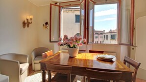 A dining area with a wooden table, chairs, a vase of flowers, and a view of buildings through open windows. - Canale - WR Apartments (Venice)
