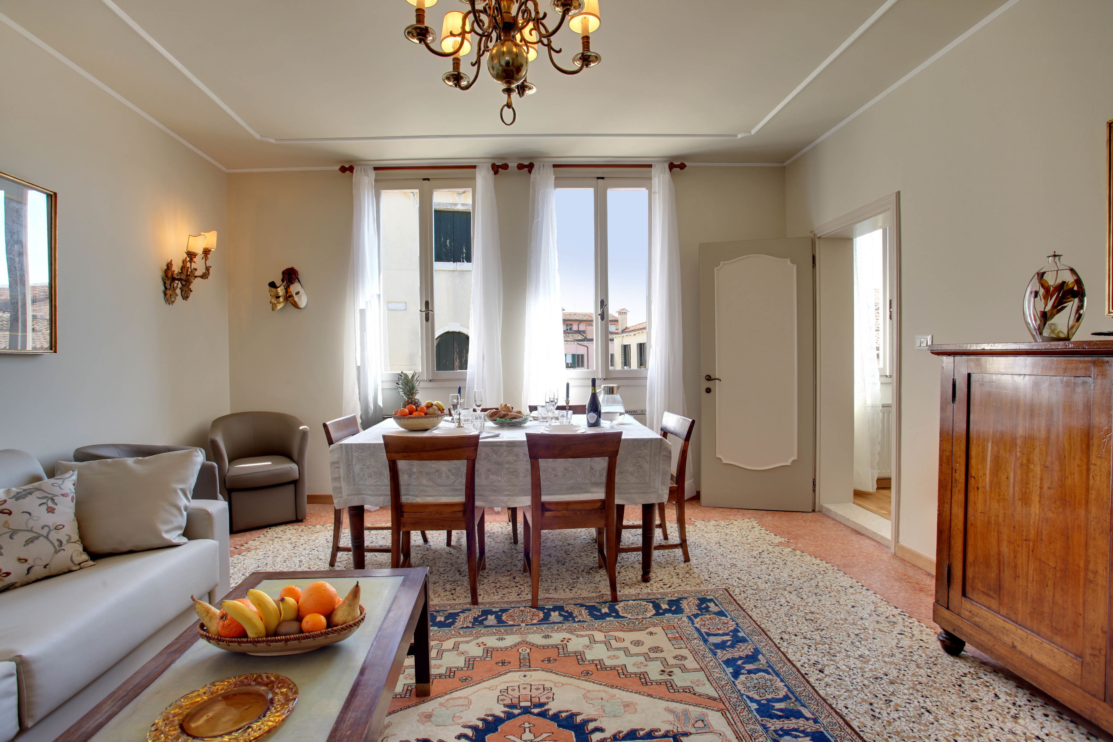 Uma sala de estar com sofá, mesa de jantar posta com frutas e copos, lustre e tapete estampado.