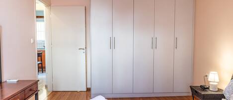 A bedroom with a bed, a desk, a wardrobe, and a nightstand with a lamp.